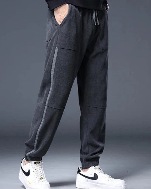 Amos Corduroy Joggers in dark grey with relaxed tapered fit, functional pockets, and ankle cuffs, styled with white sneakers.