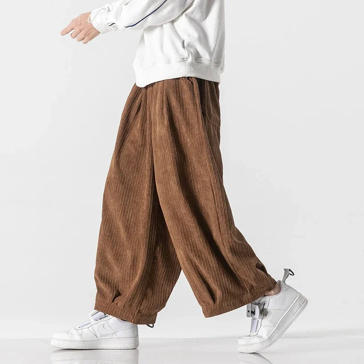 Model wearing oversized brown corduroy pants with deep pleats and elastic waistband for relaxed fit.