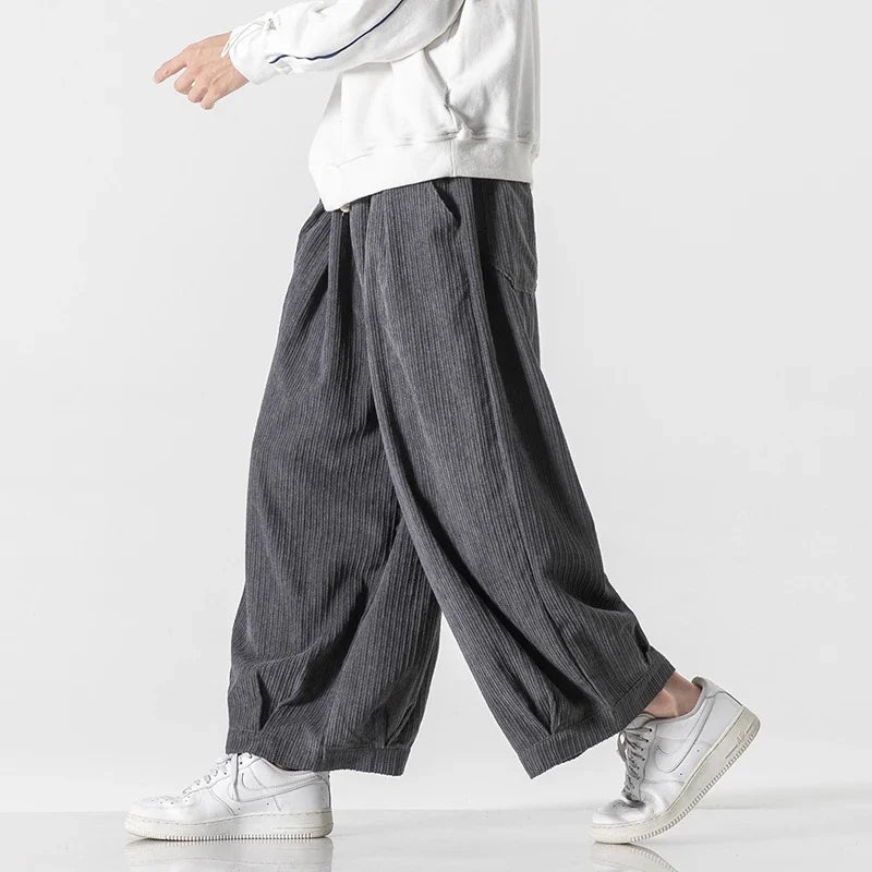 Model wearing dark oversized corduroy pants with deep pleats and relaxed fit, paired with white sneakers.
