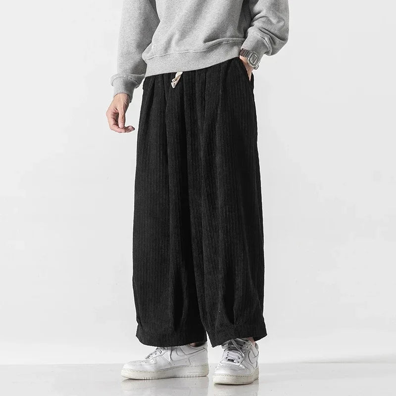 Model wearing oversized black corduroy pants with deep pleats and elastic waistband for comfortable fit.