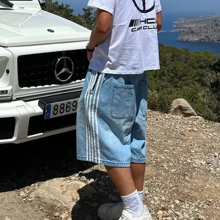 Man wearing Jax denim shorts with side stripes, standing near a luxury car in a scenic outdoor setting.