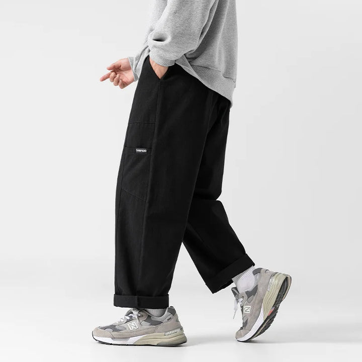 Huxley Men's 100% Cotton Cargo Pants in black with oversized pockets and adjustable drawstring waist, styled with shoes.
