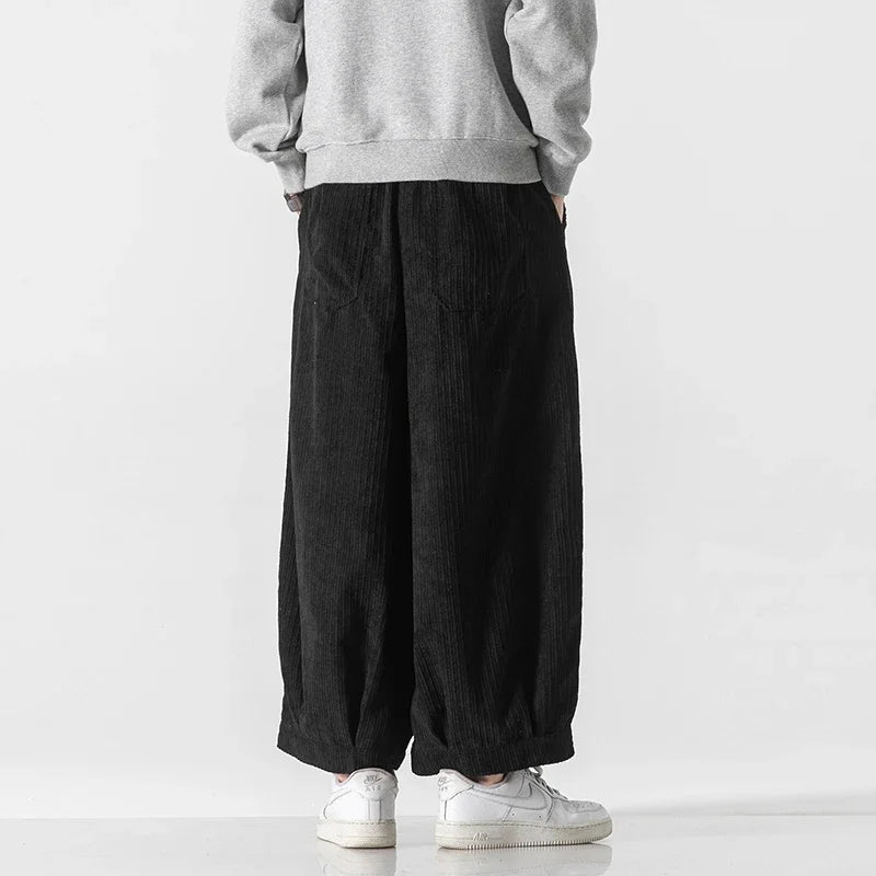 Back view of black oversized corduroy pants featuring deep pleats and a relaxed fit, paired with a gray sweatshirt.