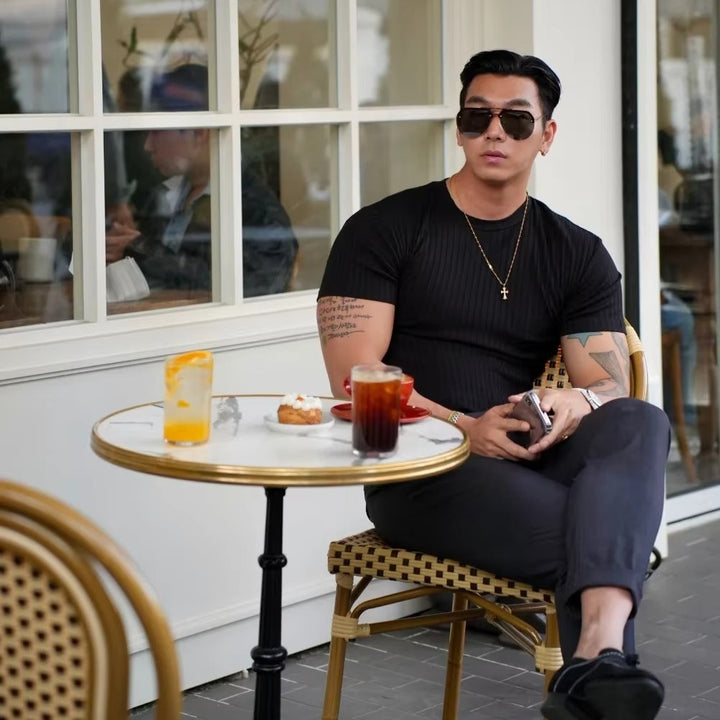 Man wearing a black ribbed knit shirt sitting at a cafe table with drinks and a pastry.