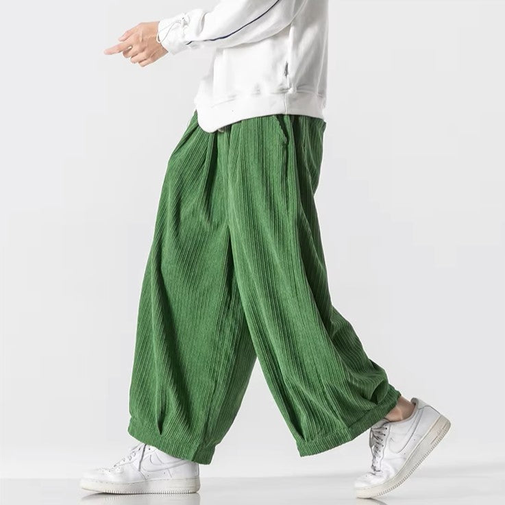 Model wearing oversized green corduroy pants with deep pleats and an elastic waistband for a relaxed fit.
