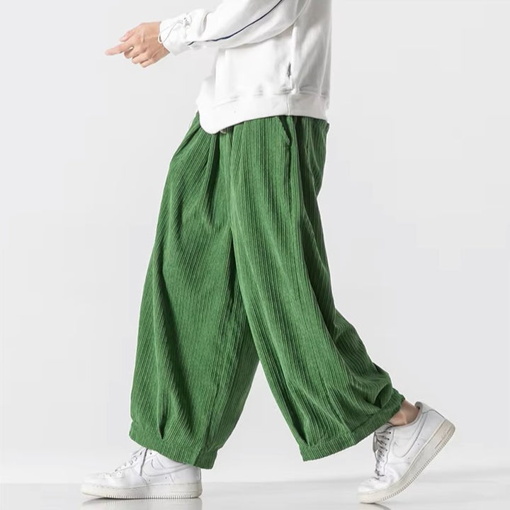 Model wearing oversized green corduroy pants with deep pleats and an elastic waistband for a relaxed fit.